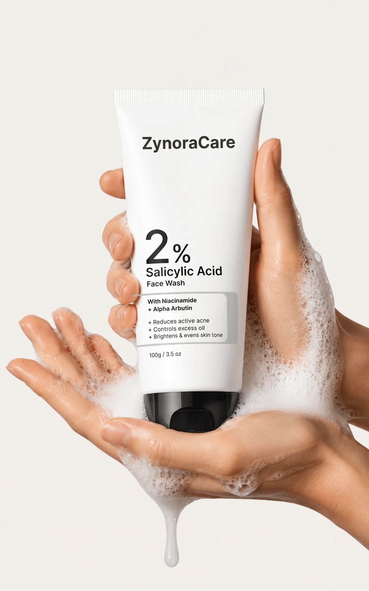 A white tube of ZynoraCare 2% Salicylic Acid Face Wash is held between two hands covered in lathered foam, against a light beige background.